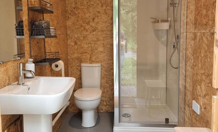 A small room with wooden walls, a shower cubicle, a toilet and wash basin with a mirror above it. There is a wire shelf on the wall